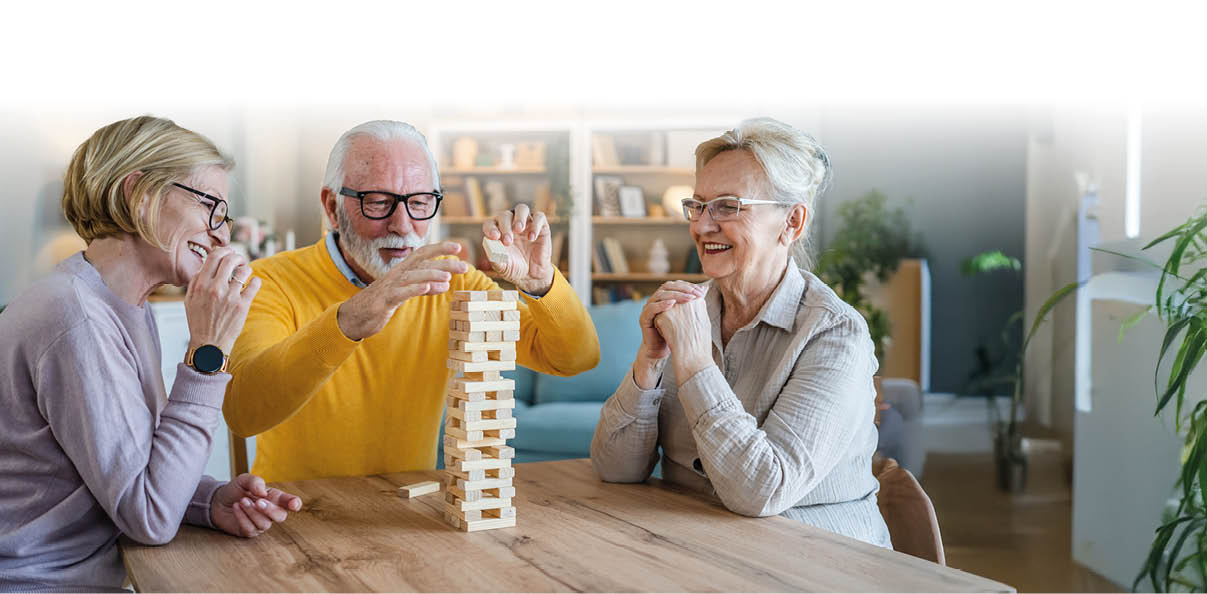 Das Bild zeigt drei ltere Menschen, die gemeinsam an einem Tisch sitzen und Jenga spielen. Alle drei wirken fr hlich und konzentriert – es wird gelacht, gestaunt und mitgefiebert. Die Szene spielt offenbar in einem hellen, gem tlichen Wohnzimmer mit Pflanzen und B cherregalen im Hintergrund.    Die Atmosph re strahlt Lebensfreude, Geselligkeit und geistige Aktivit t aus – perfekt geeignet f r Themen rund um aktive Senioren, Gemeinschaft, Freizeitgestaltung oder Gehirntraining im Alter.    