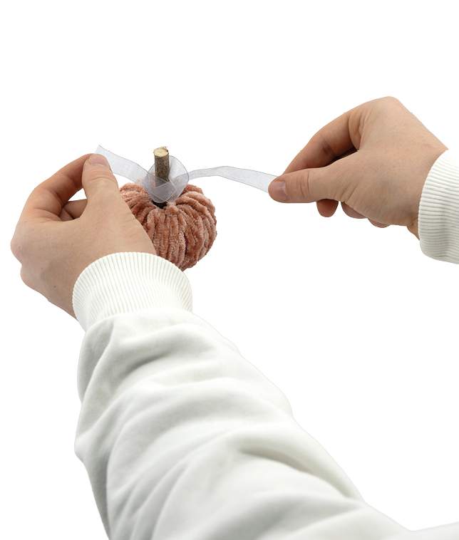 Auf dem Bild ist zu sehen, wie zwei H nde ein kleines, handgefertigtes Objekt verzieren, das wie ein K rbis aussieht. Der K rbis besteht aus flauschiger, orangefarbener Wolle, und oben steckt ein kurzer Holzstab als Stiel. Die Person bindet gerade ein durchsichtiges, helles Band (vermutlich Organza oder ein hnliches Material) um den Stiel, um dem K rbis eine dekorative Schleife zu verleihen. 