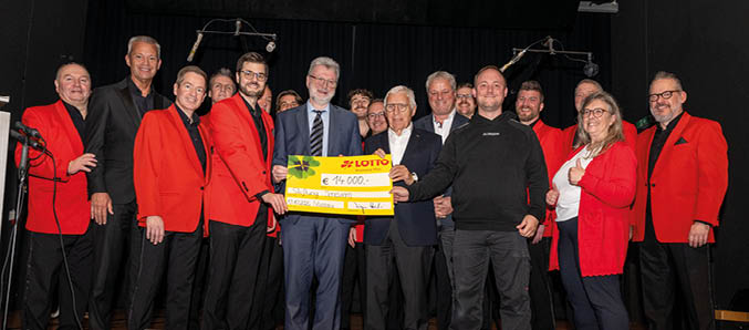 Auf dem Bild steht eine gr ere Gruppe von Menschen auf einer B hne. Mehrere Personen tragen schwarze Hosen und rote Jacken, w hrend andere in dunklen oder formellen Kleidungsst cken gekleidet sind. In der Mitte der Gruppe wird ein gro er gelber symbolischer Scheck pr sentiert. Auf dem Scheck ist ein Betrag von 14.000 Euro vermerkt. Die Personen l cheln und blicken in Richtung der Kamera. Im Hintergrund sind B hnenvorh nge und Beleuchtungseinrichtungen zu sehen..