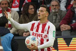 Ein junger Fu ballspieler steht am Spielfeldrand und bereitet sich darauf vor, den Ball einzuwerfen. Er tr gt das Heimtrikot des Fu ballvereins 1. FC K ln, das wei mit roten L ngsstreifen ist. Auf dem Trikot sind mehrere Sponsorenlogos zu sehen, darunter das Logo von „REWE“ in gro er roter Schrift auf der Brust sowie das gr ne „DEVK“-Logo auf dem linken  rmel. Der Spieler h lt den Ball mit beiden H nden vor sich. Sein rechter Unterarm ist bandagiert oder getaped. Er schaut nach links und scheint zu sprechen oder eine Anweisung zu geben. Im Hintergrund sitzen Zuschauer auf der Trib ne, viele davon tragen Schals in den Vereinsfarben Rot und Wei . Die Szene wirkt lebendig und spielt sich w hrend eines Fu ballspiels ab.