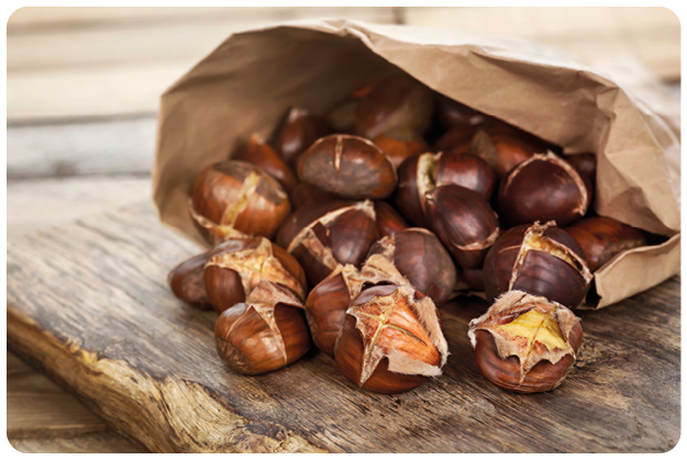 Das Bild zeigt eine Nahaufnahme von ger steten Maronen (Esskastanien), die aus einer ge ffneten braunen Papiert te auf eine rustikale Holzoberfl che herausrollen. Der Fokus liegt auf den Kastanien im Vordergrund, die warm beleuchtet sind und ihre aufgesprungenen, goldbraunen Schalen zeigen – ein Zeichen, dass sie frisch ger stet wurden.   Die Texturen sind deutlich sichtbar: die gl nzend glatte, dunkelbraune Au enschale, das leicht samtige Fruchtfleisch, das an einigen Stellen hervorlugt, sowie die raue, nat rlich gemaserte Holzplatte darunter. Der Hintergrund ist weich unscharf, wodurch die Aufmerksamkeit vollst ndig auf die Kastanien gelenkt wird.