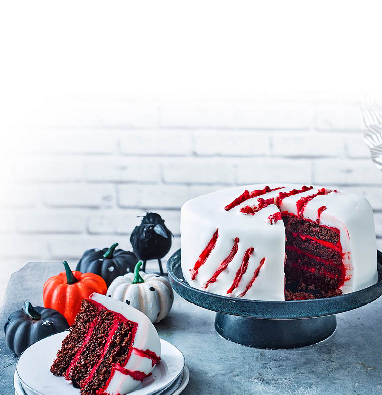 Das Bild zeigt eine mehrschichtige Torte auf einer schwarzen Tortenplatte. Die Torte ist au en mit glattem, wei em Fondant berzogen. Auf der Oberfl che und an der Seite sind rote Linien und Risse zu sehen, die an Kratzspuren erinnern und der Torte ein gruseliges Halloween-Design verleihen. Im Anschnitt erkennt man mehrere dunkle Schokoladenb den, die mit einer leuchtend roten Creme geschichtet sind. Daneben steht ein Teller mit einem St ck der Torte. Im Hintergrund sind kleine Zierk rbisse in Wei , Schwarz und Orange zu sehen – passend zur herbstlichen Halloween-Stimmung.