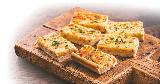  Das Bild zeigt mehrere rechteckige St cke von frisch gebackenem Zwiebelkuchen, die auf einem rustikalen Holzbrett angerichtet sind. Der Zwiebelkuchen hat eine goldgelbe, leicht gebr unte Oberfl che und ist mit fein geschnittenem Schnittlauch bestreut. Die F llung wirkt saftig und besteht sichtbar aus gehackten Zwiebeln und kleinen Speckw rfeln, die in einer cremigen Eier-Sahne-Masse eingebettet sind. Der Boden ist dick und hell, was auf einen klassischen Hefeteig hindeutet.    Die Pr sentation auf dem alten, groben Schneidebrett unterstreicht den bodenst ndigen und herzhaften Charakter dieses traditionellen Gerichts.