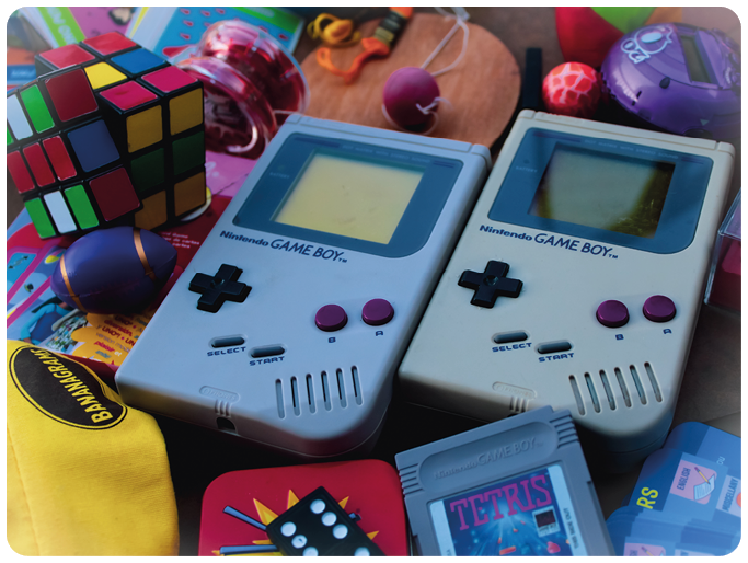 Zwei graue Nintendo Game Boy mit violetten Tasten, umgeben von Spielzeug wie Rubik's Cube, Jo-Jo und Tetris-Spielmodul.