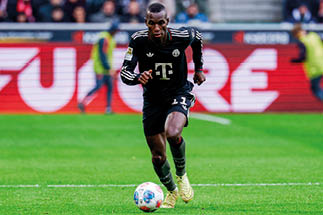 Fu ballspieler in schwarzem Trikot mit wei em Logo und gelben Schuhen l uft mit Ball auf gr nem Spielfeld.