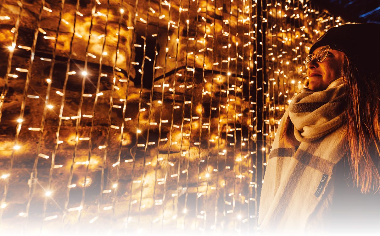 Eine junge Frau steht rechts im Bild und schaut auf einen stimmungsvollen Lichtervorhang im Hintergrund. Die Frau tr gt eine M tze und einen gro en karierten Schal.