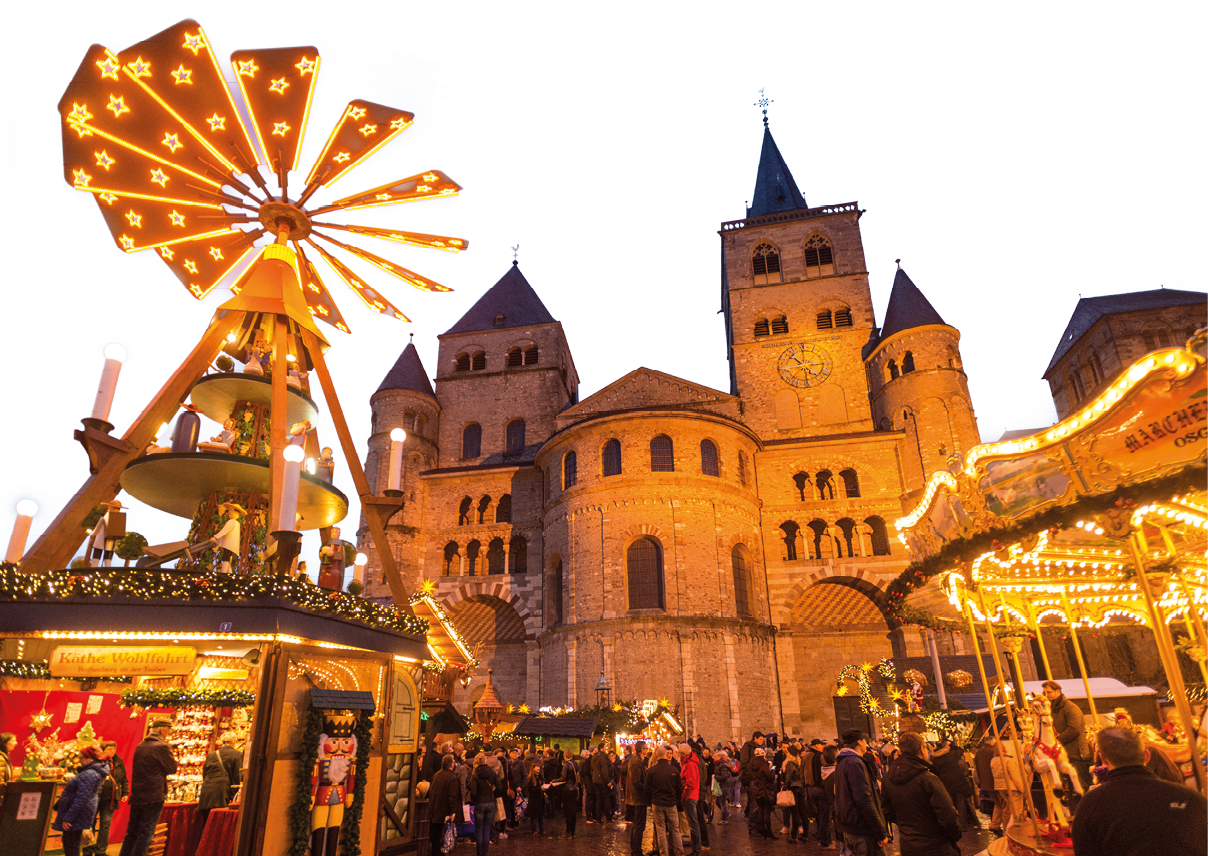 Das Bild zeigt eine stimmungsvolle Szene auf einem Weihnachtsmarkt bei Abendd mmerung. Im Vordergrund sind zahlreiche Menschen zu sehen, die ber den Markt schlendern.    Links steht eine gro e, hell erleuchtete Weihnachtspyramide mit mehreren Etagen und Figuren, die sich unter der beleuchteten Fl gelradkonstruktion drehen. Im Hintergrund ist eine Kirche zu erkennen.    Die warme Beleuchtung, das bunte Treiben und die winterliche Atmosph re erzeugen ein typisches, gem tliches Weihnachtsmarktgef hl.