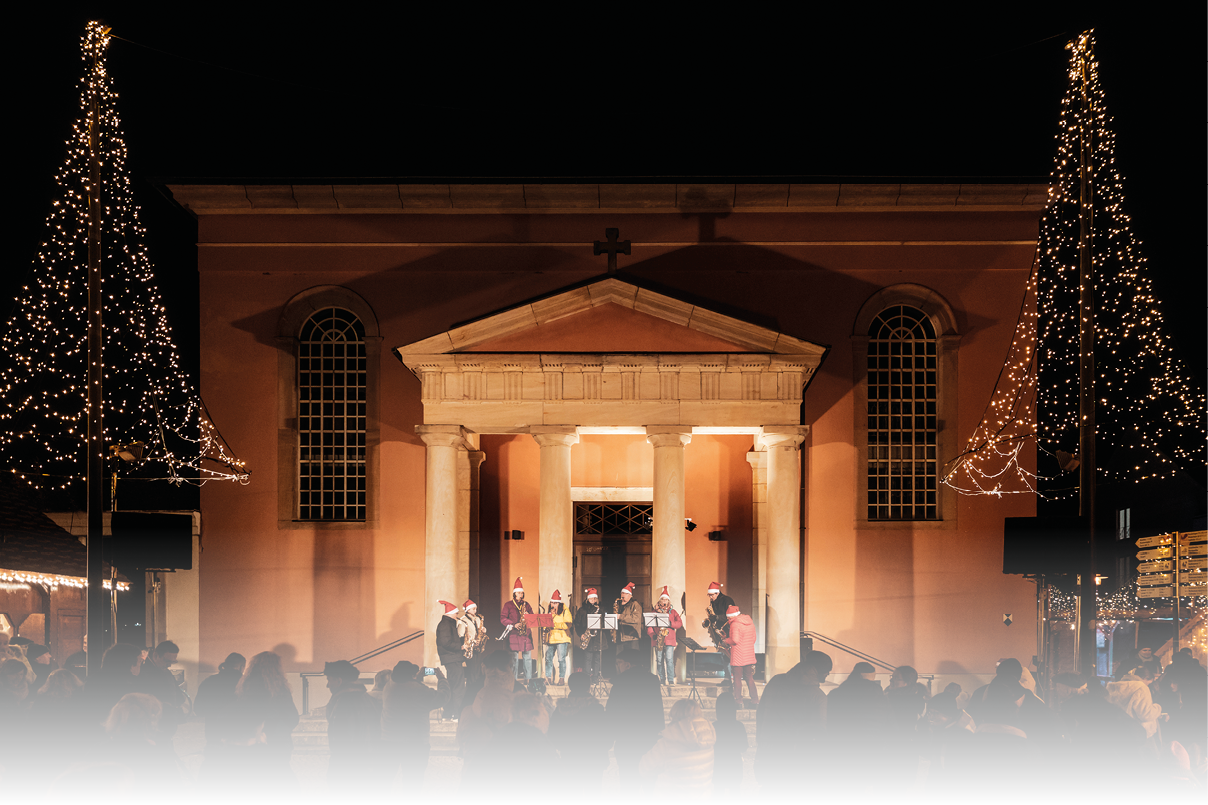 Vor der festlich beleuchteten Kirche versammelten sich am Abend zahlreiche Besucherinnen und Besucher, um die stimmungsvolle Atmosph re zu genie en. Zwei geschm ckte Weihnachtsb ume mit funkelnden Lichterketten rahmten die Szenerie ein und tauchten den Platz in warmes Licht. Auf den Stufen vor dem Kircheneingang trat eine kleine Musikgruppe auf und sorgte mit weihnachtlichen Kl ngen f r besinnliche Stimmung. Das Zusammenspiel aus Musik, Lichtern und der historischen Kulisse verlieh dem Moment eine besonders festliche und friedliche Atmosph re – ein stimmungsvoller Auftakt in die Adventszeit.