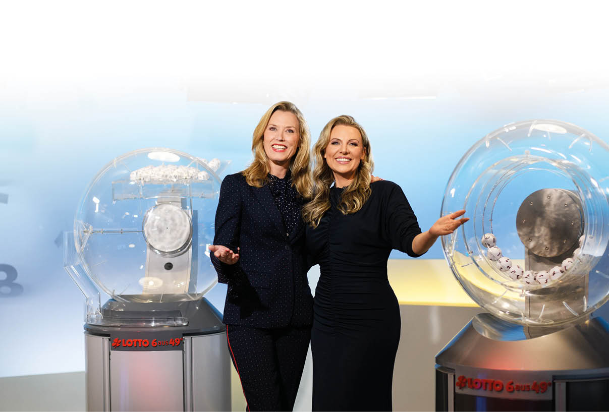 Das Bild zeigt zwei Frauen, die in einem Fernsehstudio zwischen zwei Lotto-Ziehungsmaschinen stehen. Beide l cheln freundlich in die Kamera. Auf den Maschinen ist das Logo „LOTTO 6 aus 49“ zu sehen. Die Frau links ist Franziska Reichenbacher und tr gt einen dunkelblauen Hosenanzug mit feinem Muster, die Frau rechts ist Miriam Hannah und tr gt ein elegantes, dunkelblaues Kleid. Im Hintergrund ist eine Studiowand in Blau- und Gelbt nen zu erkennen, beleuchtet von Scheinwerfern ber ihnen.