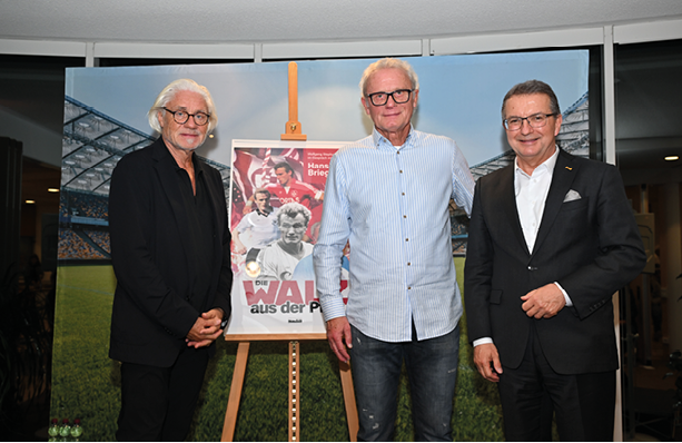 Das Bild zeigt drei Lotto-Gesch ftsf hrer J rgen H fner, Hans-Peter Briegel und Biograf Wolfgang Stephan, die nebeneinander vor einem Poster stehen, das auf einer Staffelei pr sentiert wird. Das Poster zeigt Fu ballmotive und den Titel „Die Walz aus der Pfalz“, ein Buchcover ber den bekannten Fu ballspieler Hans-Peter Briegel. Der Hintergrund ist mit einem Stadionmotiv gestaltet, was den sportlichen Kontext unterstreicht. Die drei M nner tragen elegante bis gehobene Freizeitkleidung – zwei von ihnen im Anzug oder Sakko, einer in einem hellblauen Hemd und Jeans. Sie l cheln in die Kamera.