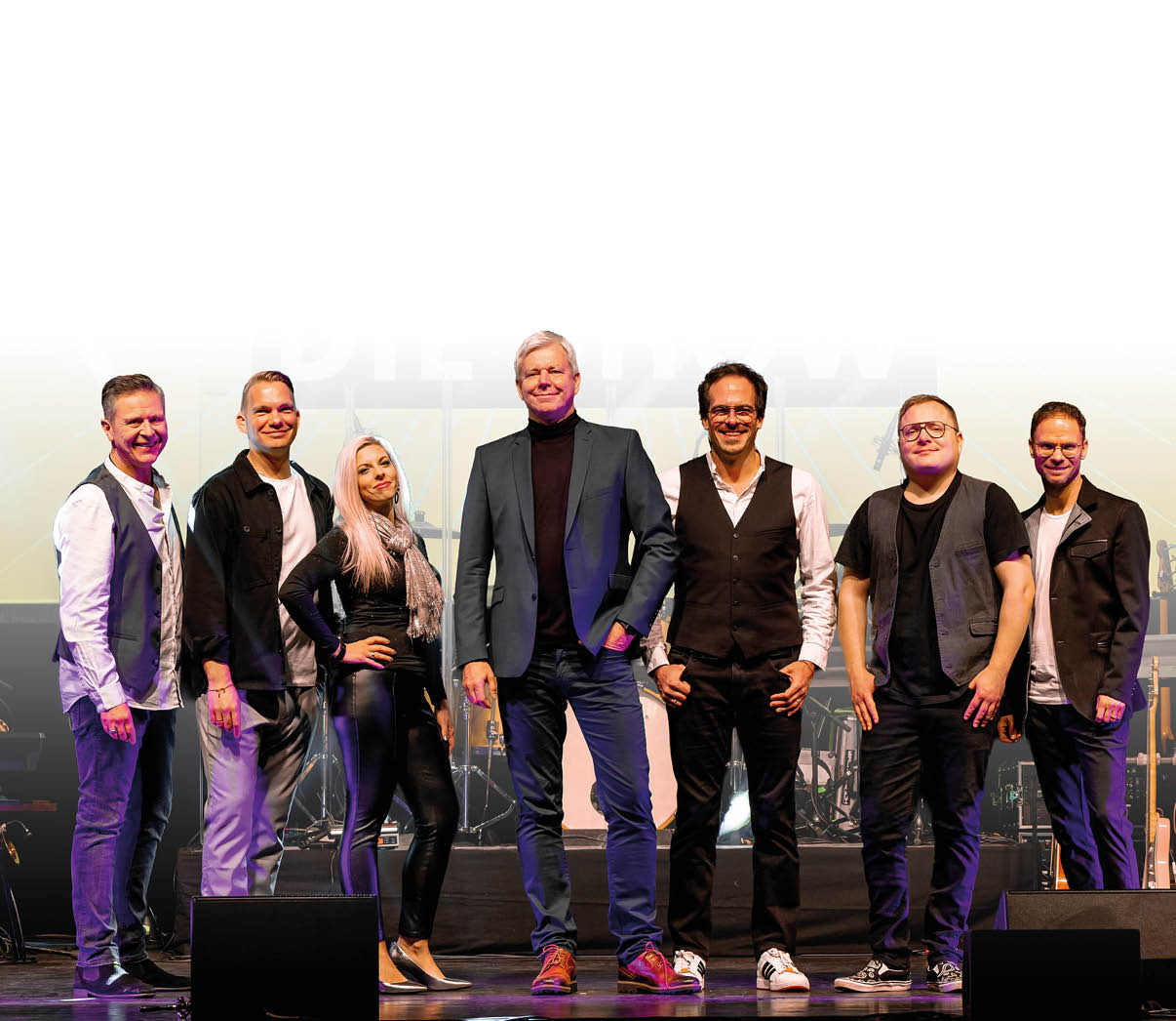 Eine bestens gelaunte Musikgruppe posiert gemeinsam f r das Foto: Sieben Mitglieder stehen nebeneinander, leger und stilvoll gekleidet. Die Atmosph re wirkt professionell und zugleich sympathisch – ein eingespieltes Team, das sichtlich Freude an seiner Musik hat. Im Hintergrund deuten B hnenlichter und Instrumente auf eine bevorstehende oder gerade beendete Live-Performance hin. Das Bild strahlt Energie, Zusammenhalt und Vorfreude auf das n chste musikalische Highlight aus.