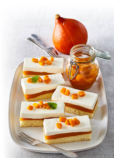 Das Bild zeigt ein herbstliches Dessert – K rbis-Schnitten mit Creme-Schicht. Auf einer rechteckigen Platte liegen mehrere St cke eines feinen Kuchens, bestehend aus drei Schichten:      einem hellen Biskuit- oder R hrteigboden,      einer leuchtend orangefarbenen K rbiscreme,      und einer dicken Schicht heller Sahne- oder Frischk secreme.      Obenauf sind kleine W rfel aus kandiertem oder eingekochtem K rbis sowie etwas Zitronenmelisse als Garnitur verteilt.  Daneben steht ein Glas mit K rbisst ckchen in Sirup – vermutlich selbst eingekocht.  Im Hintergrund liegt ein ganzer Hokkaidok rbis, der farblich perfekt zu den Dessertst cken passt.  Die Komposition wirkt appetitlich, saisonal und elegant angerichtet – ideal f r ein herbstliches Kaffeetrinken oder ein feines Buffet.