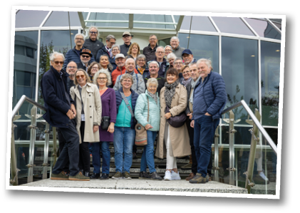 Das Bild zeigt eine Gruppe von etwa 30 Personen, die gemeinsam auf einer Treppe vor einem gl sernen Geb ude posieren. Die Gruppe besteht berwiegend aus  lteren Erwachsenen, die fr hlich in die Kamera l cheln. Viele tragen wetterfeste Kleidung wie Jacken, M ntel und Schals – es scheint ein k hler, aber heller Tag zu sein. Die Stimmung wirkt freundlich und kameradschaftlich.