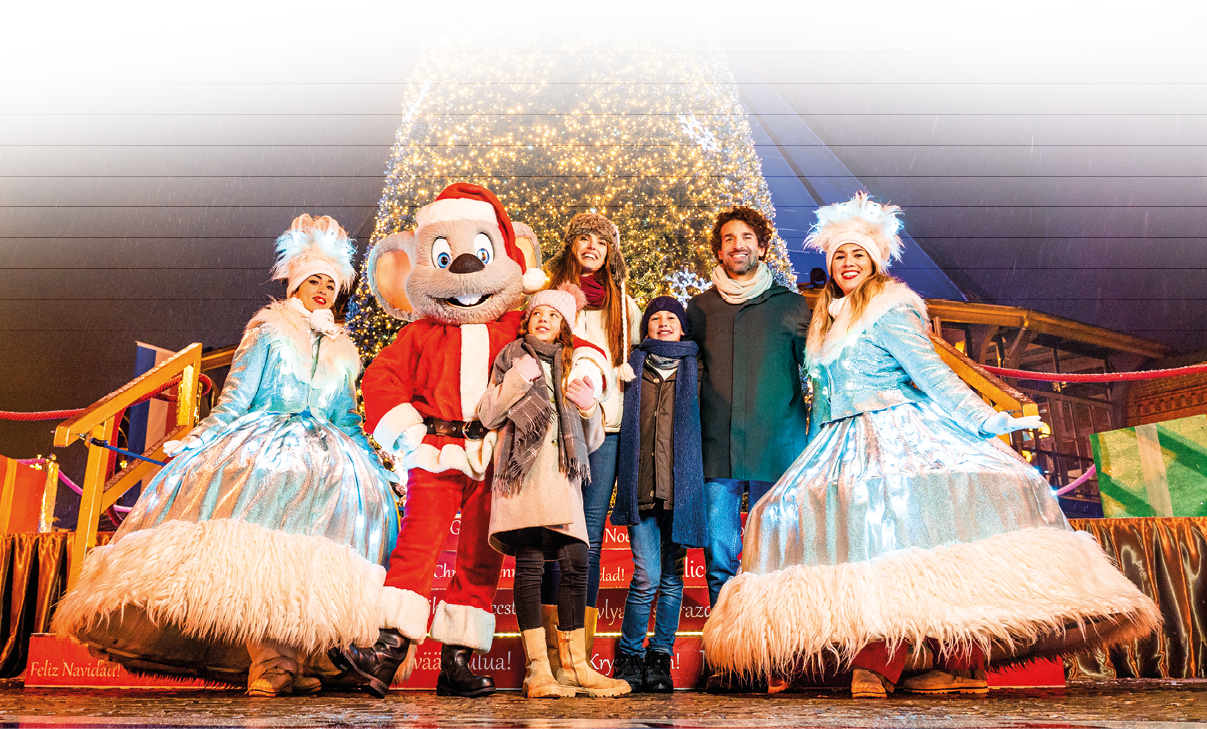 Das Bild zeigt eine fr hliche Winterszene vor einem sehr gro en, reich beleuchteten Weihnachtsbaum. In der Mitte steht eine gro e Maus-Maskottchenfigur im Weihnachtsmannkost m. Links und rechts davon posieren zwei Darstellerinnen in funkelnden, silbrig-blauen, weit ausgestellten Winterkleidern mit wei en Fellbes tzen und gro en, flauschigen Kopfbedeckungen.   Vor und neben dem Maskottchen steht eine Familie: zwei Erwachsene und zwei Kinder, warm in Winterm ntel, M tzen und Schals gekleidet. Alle l cheln und wirken begeistert. Im Hintergrund leuchten bunte Geschenkpakete und festliche Dekorationen, und es scheint leicht zu schneien. Der Boden ist nass und reflektiert die hellen Lichter der Umgebung.
