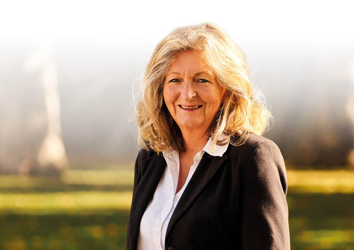 Frau mit schulterlangem, gewelltem, blondem Haar tr gt wei e Bluse und schwarzen Blazer, steht im Freien vor unscharfem Hintergrund mit B umen.