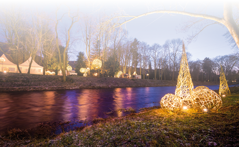Leuchtende Kugeln und kegelf rmige Lichterdekorationen am Ufer eines Flusses bei D mmerung, B ume im Hintergrund.
