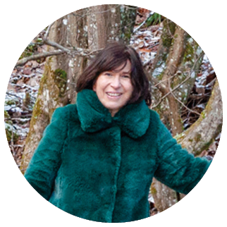 In einem winterlichen Wald steht eine Person zwischen mehreren schlanken B umen. Die Person tr gt eine gr ne, flauschige Winterjacke, einen dunkelgr nen Rock, schwarze Leggings und schwarze Winterstiefel. Sie tr gt au erdem schwarze Handschuhe und ber hrt mit einer Hand den Stamm eines Baumes. Die Person l chelt und blickt in Richtung Kamera.   Der Waldboden ist mit einer d nnen Schicht Schnee oder Frost bedeckt, sodass das Laub leicht wei   berhaucht erscheint. Hinter der Person stehen mehrere B ume und Str ucher, teils mit Restbl ttern in warmen Braun- und Oranget nen. Rechts im Bild liegt ein gro er, abgeschnittener Baumstumpf, daneben liegt Totholz.    Die Szene wirkt freundlich und ruhig und zeigt einen winterlichen Spaziergang im Wald.