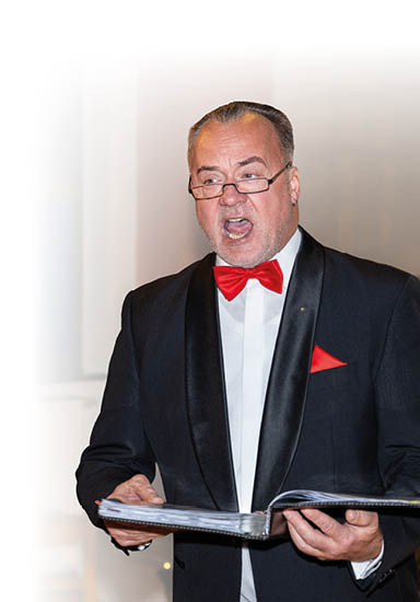 Auf dem Foto ist ein festlich gekleideter Mann der Mainzer Hofs nger zu sehen, der offenbar mitten im Gesang steht. Er tr gt einen schwarzen Smoking mit seidig gl nzendem Revers, ein wei es Hemd und eine auff llige rote Fliege, passend zum roten Einstecktuch. In den H nden h lt er eine ge ffnete Mappe, vermutlich mit Notenbl ttern. Sein konzentrierter, ausdrucksstarker Gesichtsausdruck deutet auf einen emotionalen Moment w hrend einer musikalischen Darbietung hin.