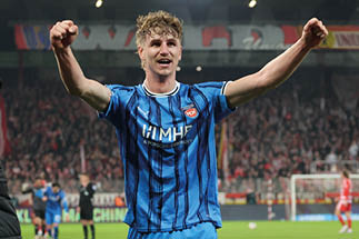 Fu ballspieler in blau-schwarzem Trikot mit erhobenen Armen, im Hintergrund Stadion mit Zuschauern und weiteren Spielern.