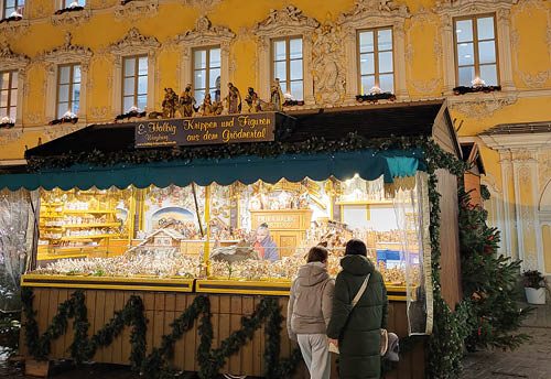 Auf dem Bild ist ein weihnachtlich dekorierter Verkaufsstand auf einem Weihnachtsmarkt zu sehen. Der Stand ist ppig beleuchtet und reich geschm ckt – mit Tannengirlanden, Lichterketten und einem dunkelgr nen Baldachin.    Im Inneren des Standes sitzt eine Person, die offenbar Krippenfiguren und Krippenzubeh r verkauft.  ber dem Stand h ngt ein Schild mit der Aufschrift:    „Krippen und Figuren aus dem Gr dnertal“    Zwei Besucherinnen oder Besucher stehen davor und betrachten die ausgestellten Waren, die von traditionellen Holzfiguren bis hin zu ganzen Krippenszenen reichen. Der Hintergrund zeigt ein festlich beleuchtetes Geb ude, was die warme, weihnachtliche Atmosph re noch verst rkt.