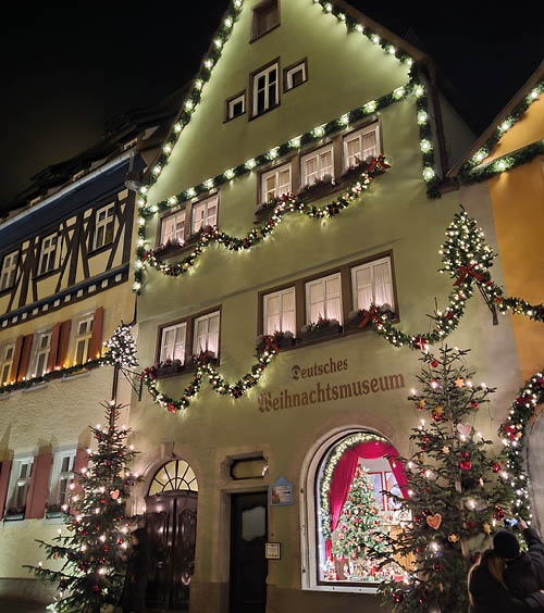 Das Foto zeigt eine festlich beleuchtete Hausfassade in traditioneller Fachwerkarchitektur – es handelt sich um das Deutsche Weihnachtsmuseum. Die Fassade ist reich mit Lichterketten, Girlanden und roten Schleifen geschm ckt. In den Fenstern stehen Weihnachtsschmuck und Kerzendekorationen, die warmes Licht nach au en werfen.   Rechts und links vor dem Geb ude stehen geschm ckte Weihnachtsb ume, die mit Kugeln, Lichtern und weiteren Ornamenten dekoriert sind. Der Eingang ist einladend erleuchtet, im Inneren ist bereits ein gro er Weihnachtsbaum zu erkennen. Die gesamte Szene wirkt sehr stimmungsvoll und typisch vorweihnachtlich – voller traditionellem Charme und festlicher Atmosph re.