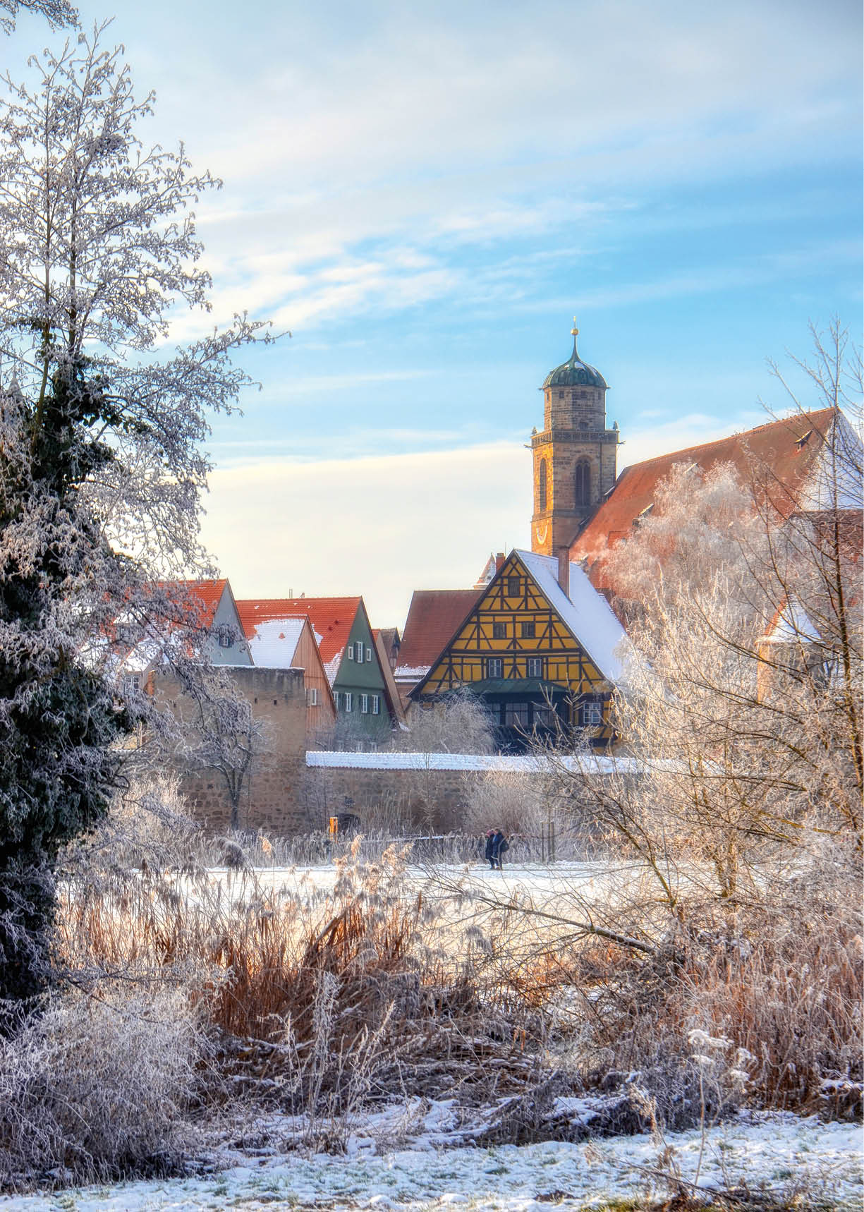 Das Bild zeigt eine malerische Winterstadt-Szenerie in typisch s ddeutscher Bauweise. Besonders auff llig sind:   Historische Altstadt mit Fachwerkh usern  Im Mittelpunkt steht ein pr chtiges gelb-schwarzes Fachwerkhaus, eingerahmt von weiteren historischen Geb uden mit roten D chern. Diese Architektur ist typisch f r St dte in Franken oder im s ddeutschen Raum.    Kirchturm als markantes Wahrzeichen  Der gro e, sandsteinfarbene Kirchturm mit gr nlicher Kuppel sticht hervor – ein Hinweis auf eine bedeutende Stadtkirche. Der Stil passt gut zu mittelalterlichen Stadtkernen.    Winterliche Stimmung    B ume sind von Raureif  berzogen    Schnee bedeckt D cher, Mauern und Wege    Warme Sonnenstrahlen schaffen einen weichen, fast m rchenhaften Glanz