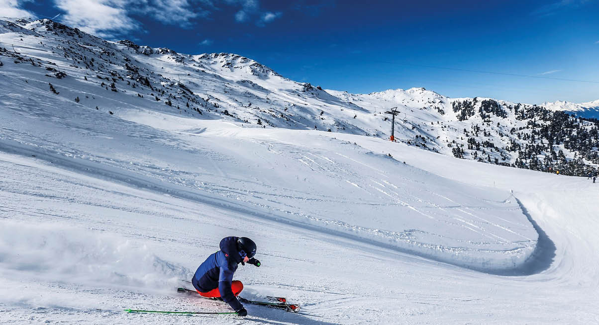 Das Bild zeigt eine Person, die auf einer schneebedeckten Piste Ski f hrt. Die Person tr gt eine blaue Jacke, rote Skihosen, schwarze Handschuhe und einen schwarzen Helm. In dynamischer Kurvenlage zieht sie eine enge Spur durch den Schnee, wobei feiner Pulverschnee zur Seite spritzt. Im Hintergrund breiten sich verschneite Berggipfel unter einem strahlend blauen Himmel aus. Die Szene vermittelt Bewegung, sportliche Energie und winterliche Frische.