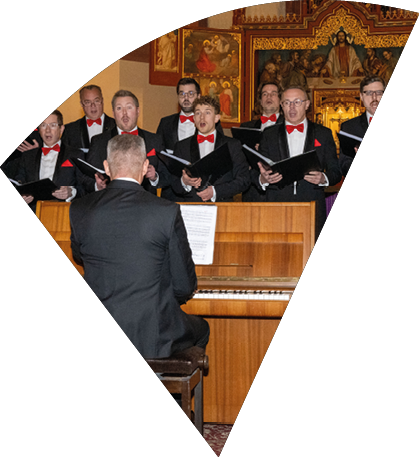 Auf dem Bild sind die Mainzer Hofs nger zu sehen. Die M nner stehen in mehreren Reihen und tragen festliche schwarze Anz ge mit wei en Hemden, roten Fliegen und roten Einsteckt chern. Sie halten ge ffnete Notenmappen in den H nden und singen, w hrend sie dem Dirigenten zugewandt sind, der im Vordergrund mit dem R cken zur Kamera steht. Im Hintergrund erkennt man Teile einer Kirche oder eines sakralen Raumes, unter anderem Gem lde und Ikonen an der Wand.