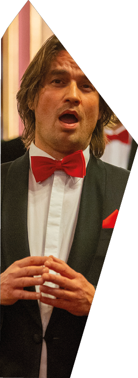 Auf dem Bild ist ein Mitglied der Mainzer Hofs nger zu sehen.   Der Mann tr gt einen schwarzen Anzug, ein wei es Hemd und eine rote Fliege, die Teil des typischen B hnenoutfits des Chors ist. Er scheint sich gerade mitten im Gesang zu befinden – sein Mund ist ge ffnet, und seine H nde sind locker vor dem K rper gefaltet. Der Bildausschnitt zeigt ihn aus der N he, sodass der Fokus klar auf seinem Gesichtsausdruck und der formellen Kleidung liegt.