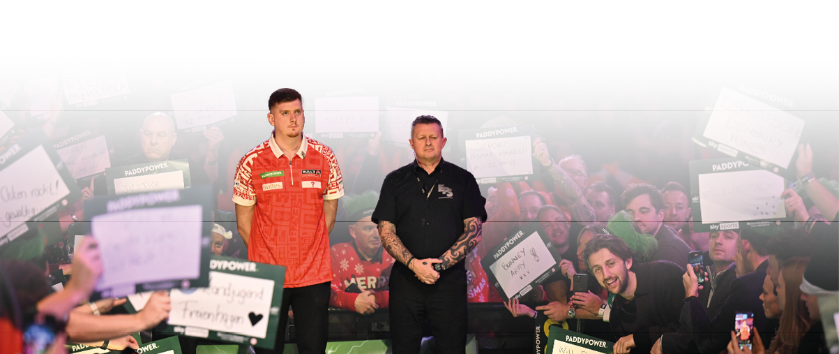 Im ber hmten “Ally Pally" stehen zwei Dartspieler auf der B hne. Der Spieler links tr gt ein rotes Shirt, der Spieler rechts ein schwarzes Shirt. Um die B hne herum stehen Fans, die Schilder hochhalten oder mit dem Handy Fotos machen.