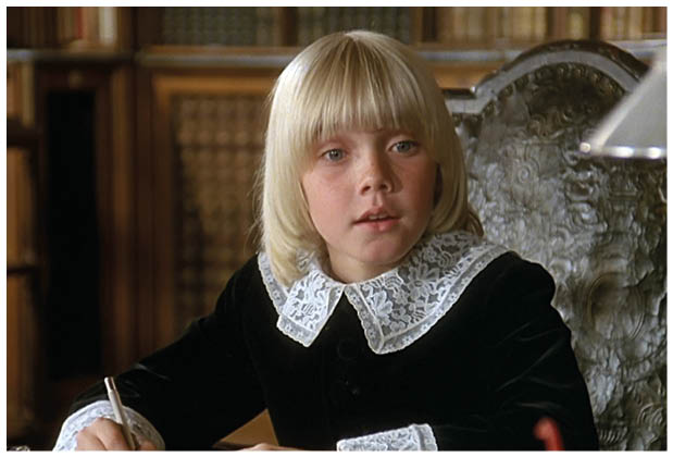 Auf dem Bild ist ein blondes Kind mit mittellangem Haar zu sehen. Die Person tr gt ein dunkles, samtartig wirkendes Oberteil mit einem gro en wei en Spitzenkragen, was dem Outfit einen historischen oder festlichen Charakter verleiht. Das Kind sitzt an einem Tisch oder Schreibtisch; im Hintergrund erkennt man eine holzget felte Wand oder ein M belst ck, das ebenfalls eher klassisch wirkt. Es ist eine Filmszene aus “Der kleine Lord".