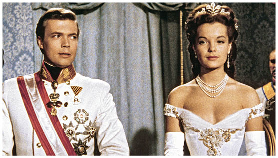 Auf dem Bild sind zwei Personen in aufwendig gestalteten historischen Kost men zu sehen.   Links steht ein Mann in einer reich verzierten wei en Uniform mit Schulterst cken, Orden und einer Sch rpe.    Rechts steht eine Frau in einem eleganten wei en Ballkleid mit tiefem Ausschnitt, Handschuhen und Schmuck, darunter eine mehrreihige Perlenkette und ein Diadem.    Beide wirken sehr f rmlich und repr sentieren offenbar eine h fische oder kaiserliche Szene in einem Prunksaal, erkennbar an den schweren Vorh ngen und den ornamentalen Tapeten im Hintergrund. Es ist eine Szene aus dem Film “Sissi".