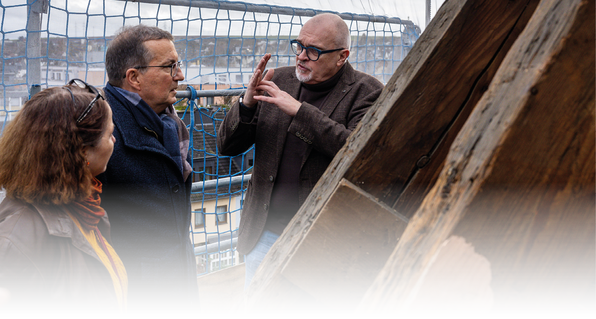 Auf dem Bild stehen drei Personen auf der Baustelle im Dachgest hl der Christuskirche in Andernach. Eine Person (rechts) erkl rt oder erl utert etwas und gestikuliert dabei mit den H nden. Die beiden anderen Personen (links) h ren aufmerksam zu. Hinter ihnen ist ein Sicherheitsnetz angebracht, wie es auf Ger sten blich ist, und Teile des Dachstuhls oder einer h lzernen Konstruktion sind deutlich sichtbar.  