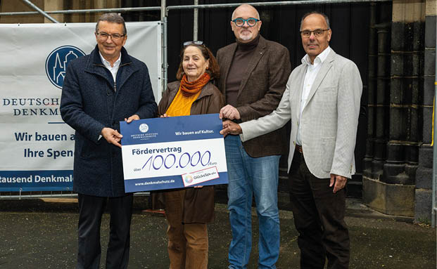 Auf dem Bild sind vier Personen zu sehen, die gemeinsam einen gro en symbolischen F rdervertrag ber 100.000 Euro pr sentieren. Die Situation wirkt offiziell und findet im Freien statt, vermutlich vor einem historischen Geb ude, das sich in Renovierung oder Restaurierung befindet – darauf weist das Banner der Deutschen Stiftung Denkmalschutz im Hintergrund hin („Wir bauen auf Kultur. Ihre Spende hilft.“).  Die Personen stehen nebeneinander, halten den Vertrag sichtbar in der Mitte und l cheln in die Kamera. Es handelt sich offenbar um eine  bergabe oder Bekanntgabe einer finanziellen F rderung zugunsten eines Denkmalschutzprojekts.