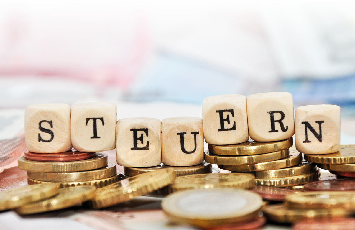 Auf dem Bild sind gestapelte M nzen in unterschiedlichen H hen zu sehen. Auf mehreren der M nzstapel liegen w rfelf rmige Holzbuchstaben, die zusammen das Wort „STEUERN“ ergeben.   Die Buchstaben sind jeweils einzeln auf den M nzen platziert und gut lesbar. Im Vorder- und Hintergrund liegen weitere lose M nzen, teils unscharf dargestellt.