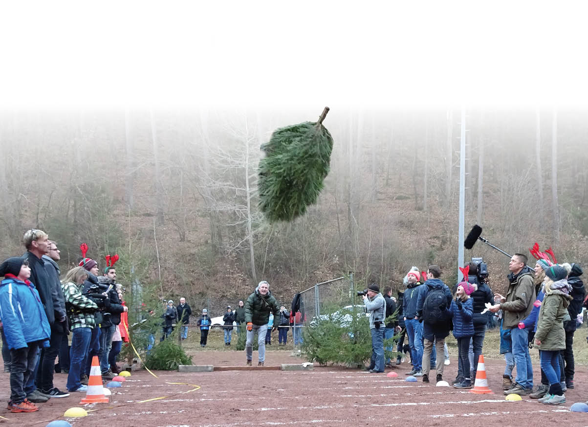 Auf dem Bild ist eine Freiluftszene mit mehreren Personen zu sehen, die sich beiderseits eines markierten Bereichs im Wald aufgestellt haben. Im Mittelpunkt befindet sich ein in die Luft geworfener Weihnachtsbaum, der sich deutlich sichtbar ber den K pfen der Anwesenden befindet.    Die Menschen stehen in zwei Reihen gegen ber, einige tragen winterliche Kleidung, M tzen oder Accessoires, vereinzelt auch rote Kopfbedeckungen. Am Boden sind Markierungen, H tchen und Ma b nder zu erkennen, was auf eine sportliche oder spielerische Wettkampfsituation hindeutet. Eine Person in der Mitte scheint den Baum soeben geworfen zu haben oder beobachtet den Wurf.    Am Rand der Szene befinden sich Zuschauerinnen und Zuschauer, darunter auch Personen mit Kamera- und Tontechnik, was darauf schlie en l sst, dass das Ereignis dokumentiert oder medial begleitet wird. Die Umgebung ist ein laubfreier Wald, was auf die Winterzeit hindeutet. Insgesamt zeigt das Bild eine organisierte Veranstaltung, bei der das Weihnachtsbaumwerfen als gemeinschaftliche Aktivit t im Vordergrund steht.