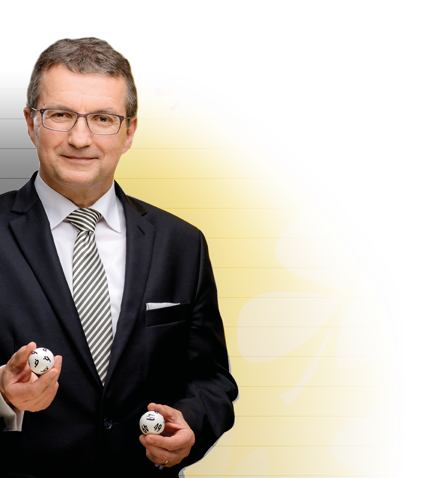 Auf dem Bild ist ein gut gekleideter Mann in Anzug und Krawatte zu sehen, der zwei wei e Lottozahlen-Kugeln in den H nden h lt. Er steht frontal zur Kamera, wirkt freundlich und seri s.