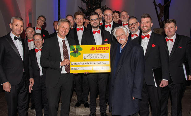 Eine Gruppe von etwa 15 M nnern steht in einer Kirche auf den Stufen vor dem Altar. Die M nner tragen berwiegend schwarze Anz ge mit wei en Hemden und roten Fliegen und geh ren zu einem M nnerchor. In der Mitte des Bildes halten zwei Personen gemeinsam einen gro en symbolischen Scheck in gelber Farbe mit der Aufschrift „Lotto Rheinland-Pfalz“ und einem Betrag von 13.000 Euro. Der Scheck ist f r soziale Zwecke bestimmt. Im Hintergrund sind Kirchenb nke, ein Altarraum sowie farbige Lichtakzente in Rot und Blau zu sehen, die eine festliche Atmosph re erzeugen. Alle Beteiligten blicken in die Kamera und wirken zufrieden und feierlich.