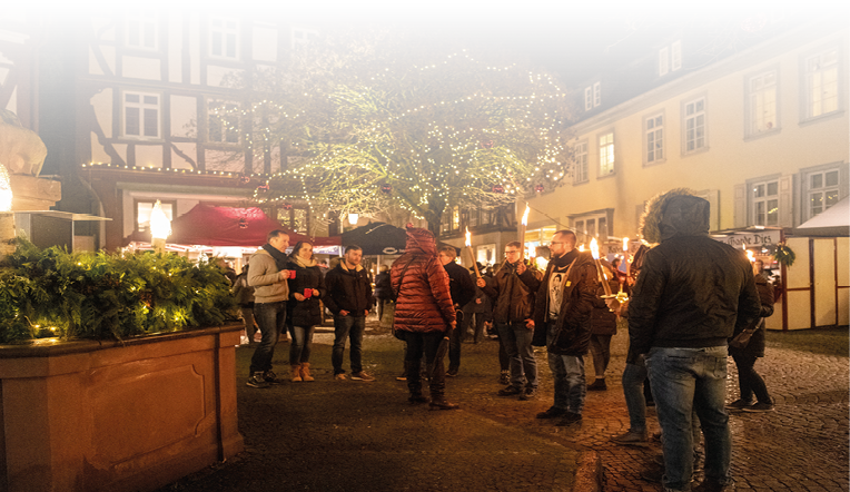 Inmitten festlich geschm ckter Fachwerkh user genie en Besucherinnen und Besucher die besondere Atmosph re des winterlichen Marktplatzes. Unter dem hell erleuchteten Baum kommen Menschen mit warmen Getr nken in der Hand zusammen, w hrend andere mit Fackeln durch die Gassen ziehen und dem historischen Ambiente zus tzlichen Zauber verleihen.