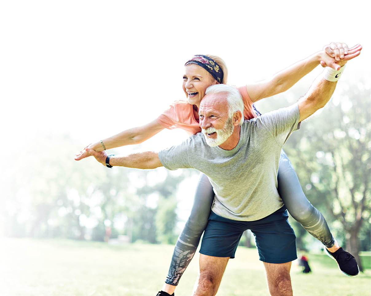 Auf dem Bild sind zwei ltere Erwachsene in einer Park- oder Gr nanlage zu sehen. Der Mann steht aufrecht und tr gt sportliche Kleidung (T-Shirt und kurze Hose). Die Frau befindet sich auf seinem R cken, h lt sich an seinen ausgestreckten Armen fest und streckt ebenfalls die Arme seitlich aus.    Beide wirken in Bewegung, stabil und ausbalanciert. Sie lachen und zeigen eine entspannte, lebensfrohe K rpersprache. Die Szene vermittelt den Eindruck von gemeinsamer sportlicher Aktivit t oder spielerischer Bewegung im Freien. Im Hintergrund sind B ume und Rasenfl chen zu erkennen, die auf eine nat rliche Umgebung hinweisen.