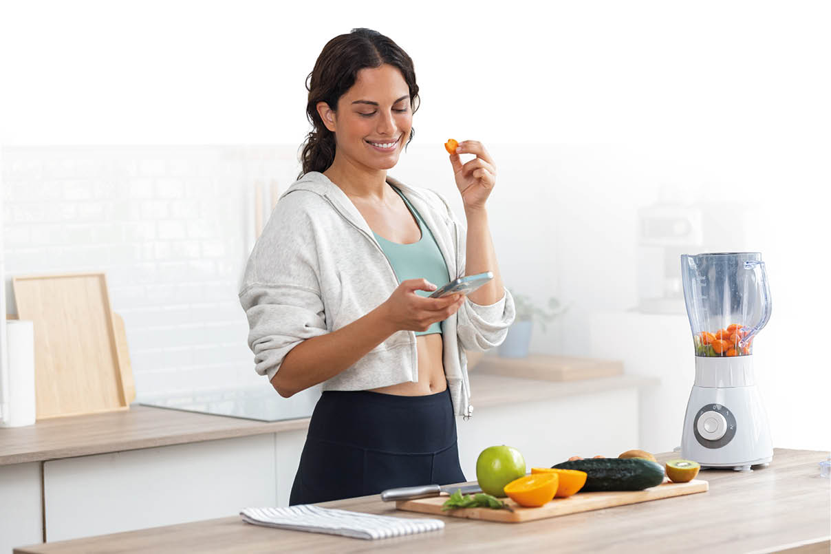 Auf dem Bild ist eine junge Frau in einer K che zu sehen. Sie tr gt sportliche Freizeitkleidung und steht an einer Arbeitsfl che, auf der frisches Obst und Gem se liegen, darunter unter anderem Orangen, ein Apfel, Gurke und Karotten.   Rechts auf der Arbeitsfl che steht ein Mixer, der bereits mit geschnittenem Gem se bef llt ist. Die Frau h lt in der einen Hand ein kleines St ck Karotte und in der anderen ein Smartphone, auf dessen Bildschirm sie blickt.    Ihr Gesichtsausdruck wirkt entspannt und zufrieden, als w rde sie eine Nachricht lesen oder ein Rezept pr fen. Die Szene vermittelt den Eindruck von Essensvorbereitung, m glicherweise f r einen Smoothie oder ein gesundes Getr nk, in einer hellen, aufger umten K chenumgebung.