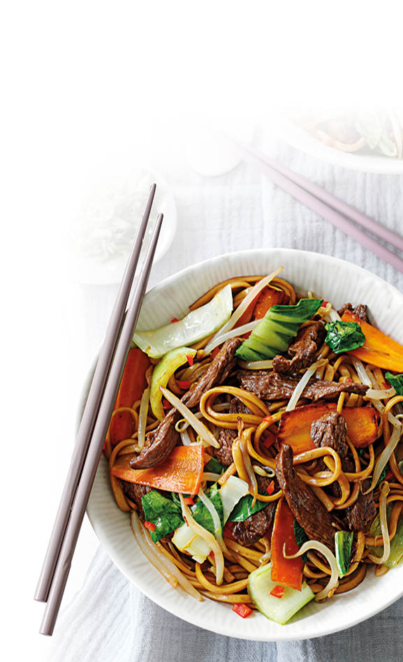 Zwei Schalen mit gebratenen Nudeln, Gem se und Fleisch, dazu zwei Paar Essst bchen und kleine Schalen mit Kr utern und Gew rzen auf grauem Stoff.