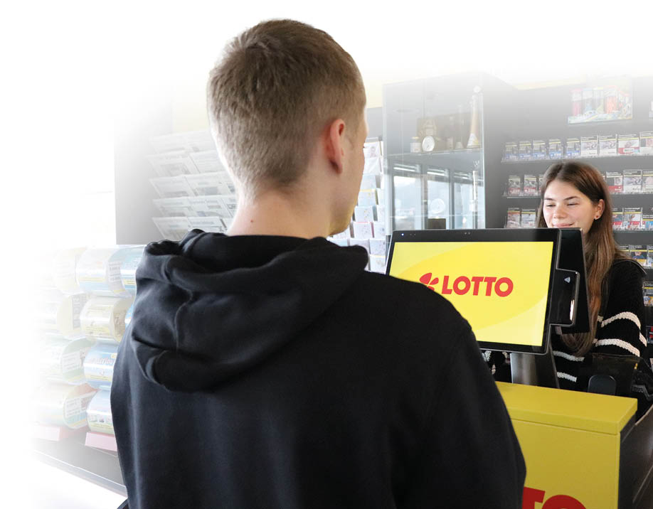 Ein junger Mann steht der Kamera abgewandt vor einem Lotto-Terminal in einem Kiosk. Hinter dem Terminal steht eine junge Frau, die das Terminal bedient. 
