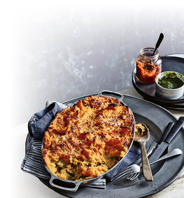 Auf einem gro en Metalltablett steht eine Auflaufform mit einem goldbraun gebackenen Auflauf, daneben liegen ein L ffel und eine Gabel, im Hintergrund stehen ein Glas mit roter Sauce und eine Schale mit gr ner Sauce.