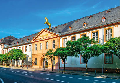 Historisches Museumsgeb ude mit einem goldenen Pferd auf dem Dachgiebel und B umen davor an einer Stra e.
