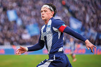 Fu ballspieler in blau-wei em Trikot mit Kapit nsbinde l uft auf dem Spielfeld, im Hintergrund unscharfe Zuschauertrib ne.