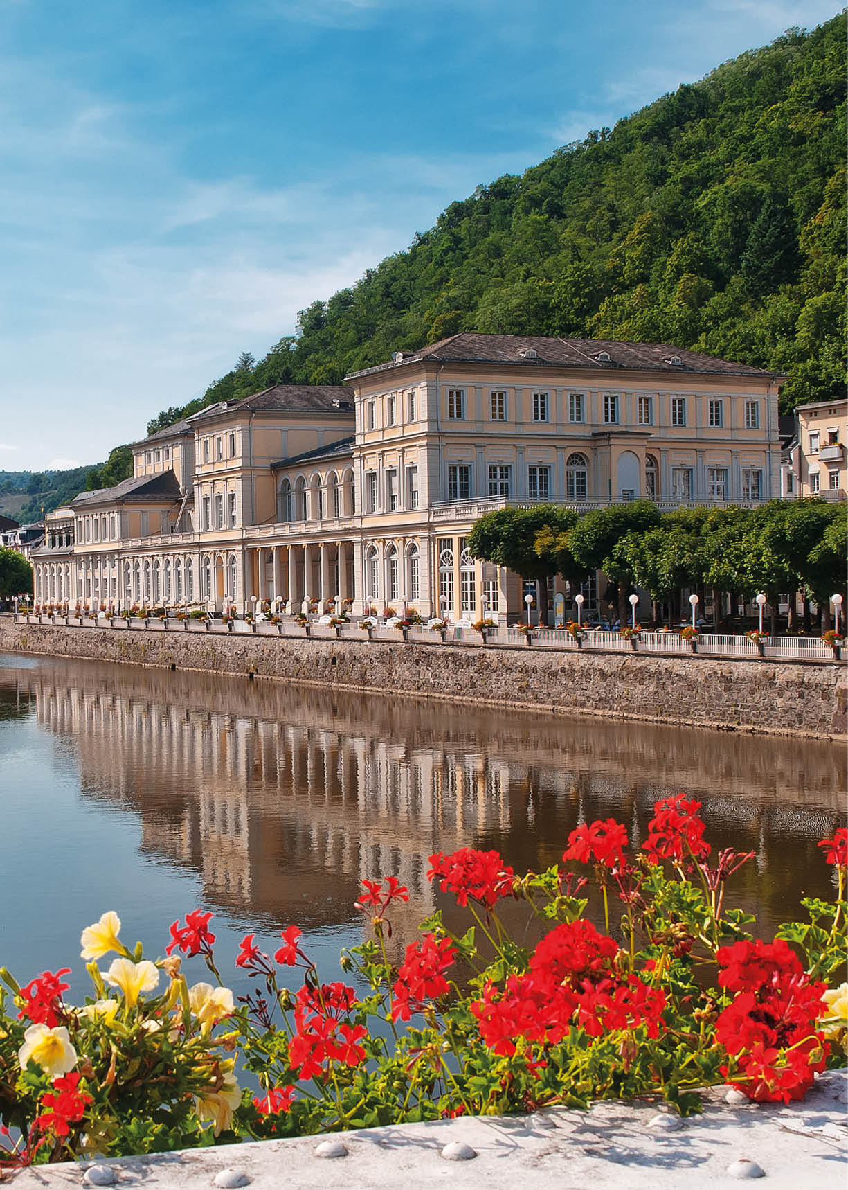 Auf dem Bild ist eine weitl ufige historische Kur- und Schlossanlage an einem Fluss zu sehen. Im Vordergrund verl uft ein ruhiges Gew sser, ber das eine filigrane Fu g ngerbr cke aus Metall mit Blumenschmuck f hrt. Am Ufer liegt ein Boot vor Anker.    Dahinter erstreckt sich ein langgezogenes, hellgelbes Geb ude mit mehreren Stockwerken, symmetrischer Fassadengliederung, vielen Fenstern und kleinen Balkonen. Das Bauwerk wirkt repr sentativ und stammt erkennbar aus dem 18. oder 19. Jahrhundert. Es handelt sich um das Kurhaus Bad Ems, das zentraler Bestandteil des historischen Kurviertels ist.    Die Anlage ist von gepflegten B umen und einer Uferpromenade umgeben. Im Hintergrund steigen bewaldete H gel steil an, was auf eine Lage in einem engen Flusstal hinweist. Der Himmel ist blau mit vereinzelten Wolken, was der Szene einen ruhigen, sommerlichen Eindruck verleiht.