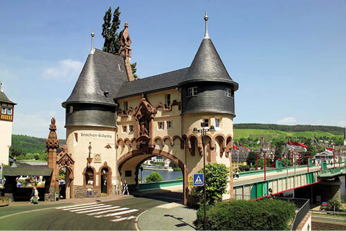 Auf dem Bild ist ein historisches Br ckentor mit zwei runden T rmen zu sehen, das als markanter Durchgang auf eine Stra enbr cke f hrt. Die T rme besitzen schiefergedeckte, spitz zulaufende D cher und sind durch einen mittleren Bauk rper mit Torbogen verbunden. In der Fassade finden sich Rundb gen, Zierelemente aus rotem Sandstein sowie eine eingelassene Figurengruppe ber dem Durchgang. Links im Geb ude ist eine Gastst tte mit der Aufschrift „Br cken-Schenke“ untergebracht.    Rechts schlie t sich die eigentliche Br cke mit Gel ndern an, die  ber einen Fluss f hrt; im Hintergrund sind weitere Geb ude eines Moselortes sowie bewaldete H nge und Weinberge zu erkennen. Das Bauwerk dient sowohl als architektonisches Wahrzeichen als auch als Zugang zur Br cke.    Es handelt sich um das Br ckentor Trarbach, eines der bekanntesten Bauwerke an der Mosel.