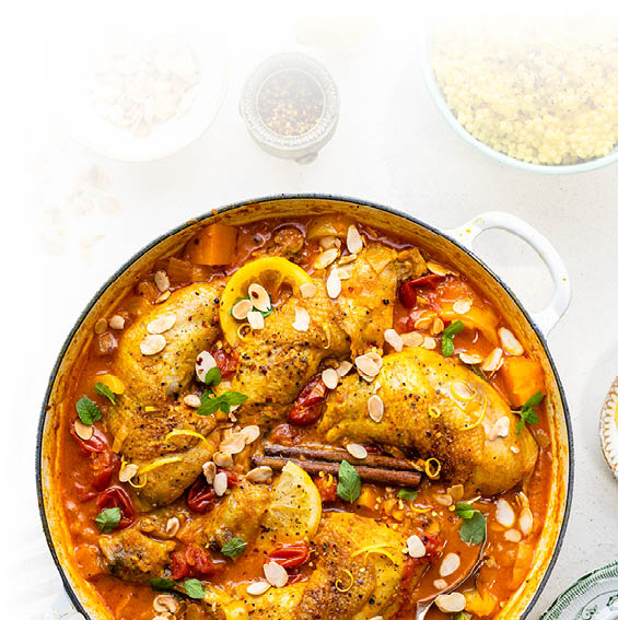 Topf mit H hnchenschenkeln in Tomatensauce, garniert mit Mandelbl ttchen, Zitronenscheiben, Zimtstangen und frischen Kr utern