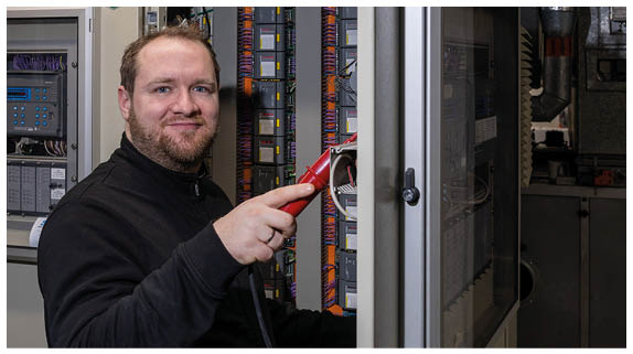 Ein Mann in dunkler Kleidung h lt ein rotes Messger t vor einem offenen Schaltschrank mit sichtbaren Kabeln und Bauteilen.